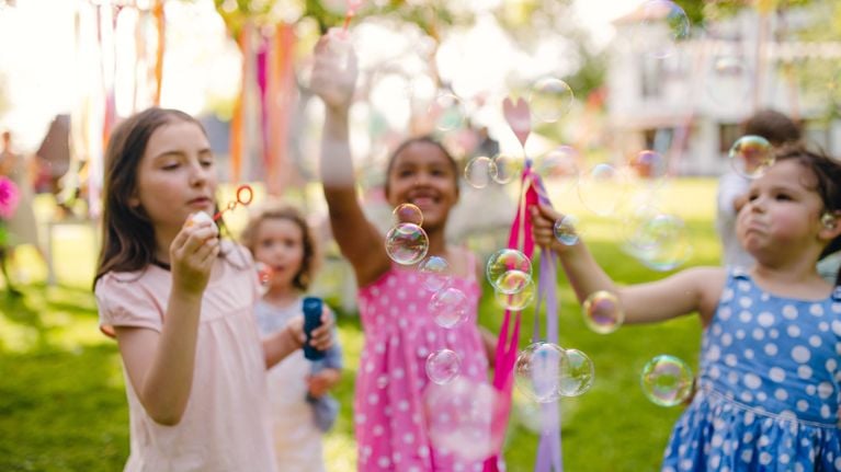 Children in bright summer clothes play with bubbles at a sunny outdoor birthday party. One girl blows bubbles in the foreground while others reach to pop them, surrounded by colorful streamers and party decorations.