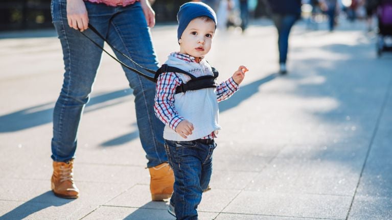 A young child in a checkered shirt, beanie, and jeans looks toward the camera while walking on a busy pedestrian path. The child is secured with a black chest-style safety harness held by an adult just behind them.