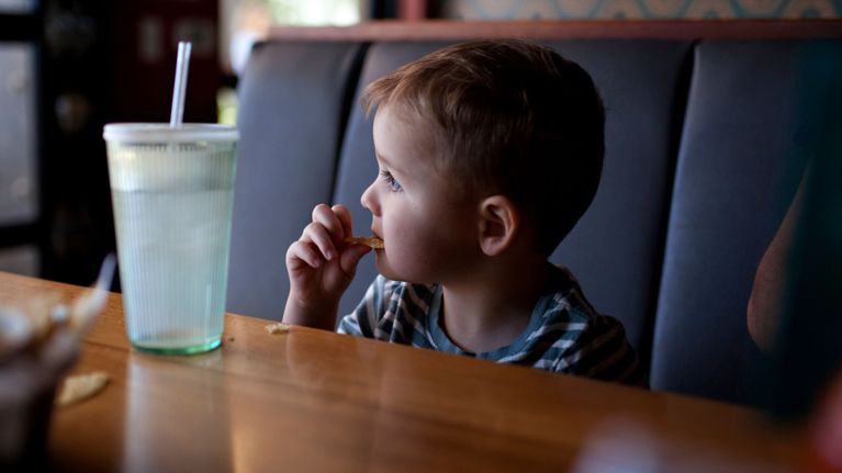 A young child sits in a restaurant booth, looking to the side while eating a chip. A large cup with a straw sits on the table in front of them. The image captures a candid moment of toddler dining, possibly in a casual family restaurant setting.