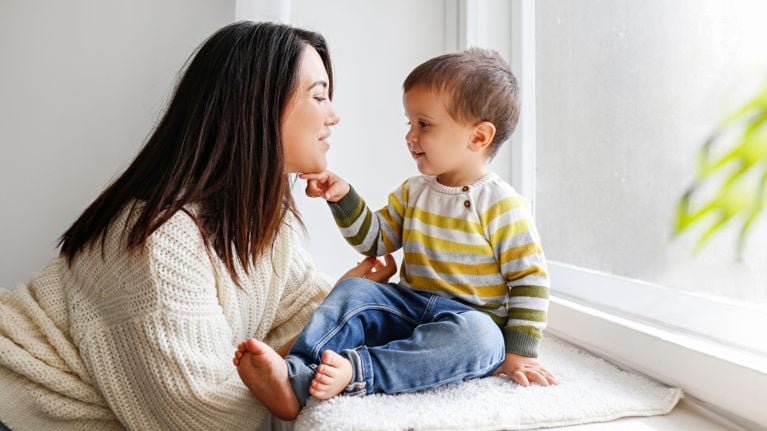 A teenage girl and her mother sit on a grey couch, smiling and making eye contact as they talk openly at home.