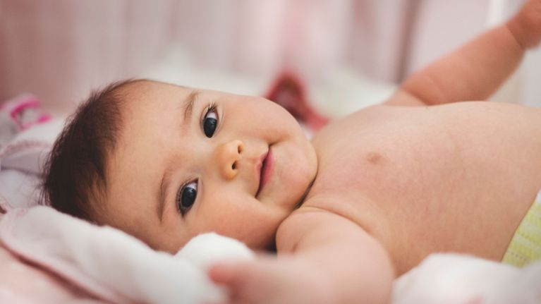 A baby lies on a soft blanket, looking at the camera with a gentle smile and outstretched arm. The baby appears relaxed and curious, showcasing the warmth and expressiveness typical of early infancy.