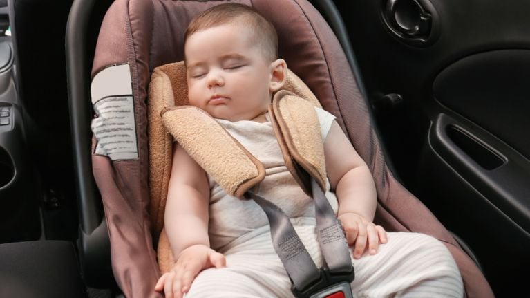 A baby peacefully sleeping in a brown infant car seat with soft beige padding, strapped in securely in the back seat of a car.