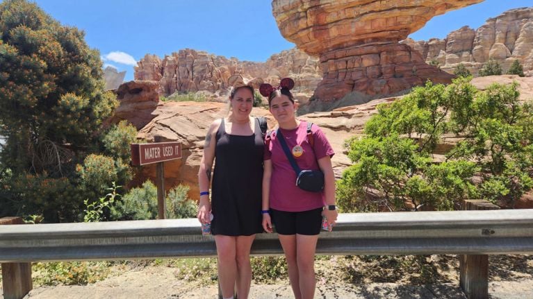 Mother and daughter wearing Mickey ears stand together in front of rocky red formations in Cars Land, beside a humorous “Mater Used It” sign.