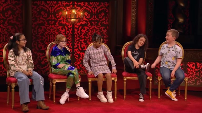 Five children sit in a row of ornate red and gold chairs against a red damask-patterned backdrop on the set of Junior Taskmaster. Each child wears bright, expressive clothing, smiling and looking engaged as they await their task or feedback.