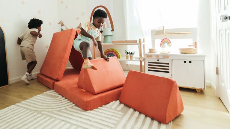 Two children play energetically on an orange Nugget couch in a bright playroom, jumping off triangular cushions and building a playful structure.