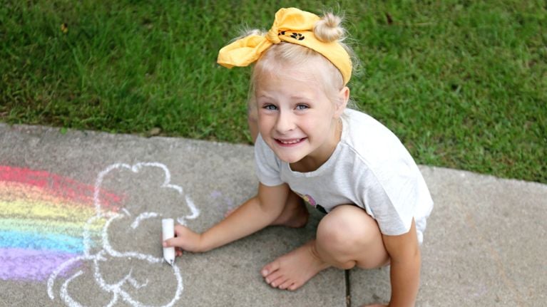 A barefoot child with a yellow bow headband draws a rainbow and cloud with sidewalk chalk on a sunny day. She looks up and smiles while crouched on the pavement near a grassy lawn.