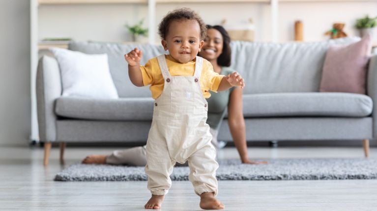 A curious toddler in a beige onesie stands in a modern kitchen, peering into an open lower drawer. The image highlights the importance of childproofing drawers and cabinets to keep toddlers safe at home.