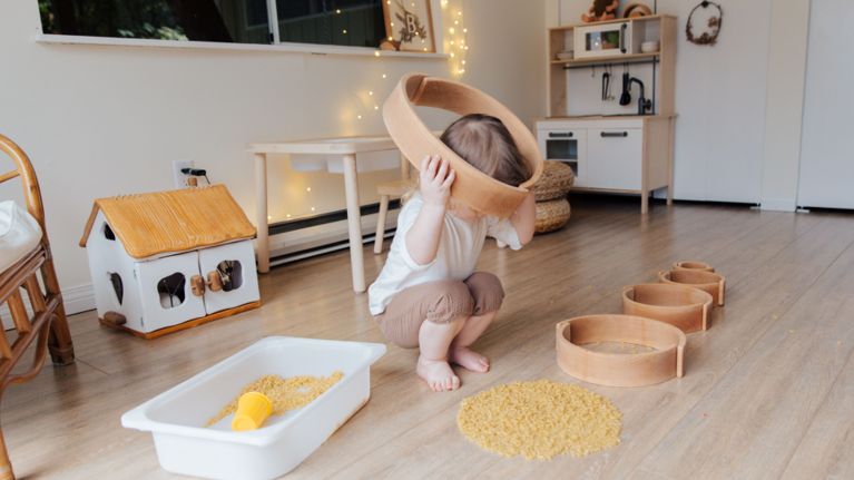 A toddler holds a wooden toy to their head, covering the eyes.