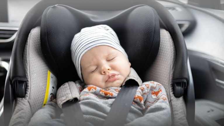 A newborn sleeping in a gray and black car seat, wearing a striped hat and a sweater with fox patterns, shown in a close-up view.