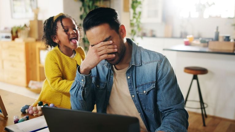 A father in a denim shirt sits at a kitchen table, head in hand, visibly overwhelmed as his young daughter playfully sticks out her tongue behind him. The image captures the tension between parental stress and childlike joy.