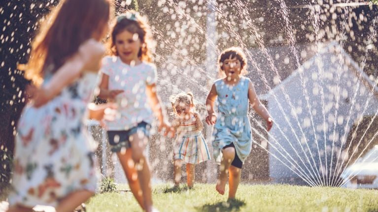 A group of children in summer clothes run and laugh through a backyard sprinkler, water droplets catching the sunlight. The scene is full of movement and joy, with blurred houses in the background.