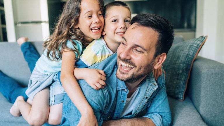 A smiling dad lies on a couch with his two young kids, who are laughing and piled on top of him. The scene radiates warmth and happiness, illustrating joyful moments of connection in fatherhood.