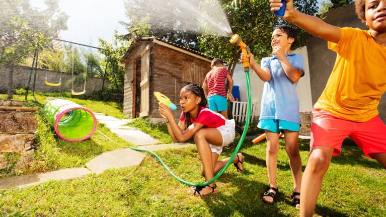 Kids engage in a lively water fight with garden hoses and water guns in a sunlit backyard. One child kneels with a water blaster while others spray water near a wooden playhouse and swings in the background.