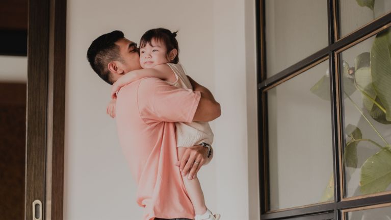 A man carries his toddler in front of a closed, paned window.