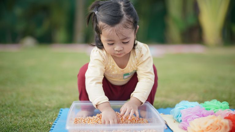 A toddler crouching in a field of grass reaches into a box.
