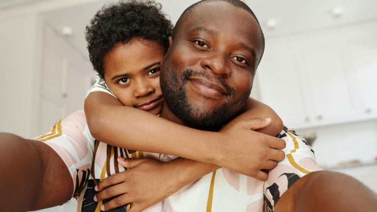 A father and child take a selfie, both looking into the camera with soft smiles. The child embraces the dad from behind, conveying love, security, and closeness in their relationship.