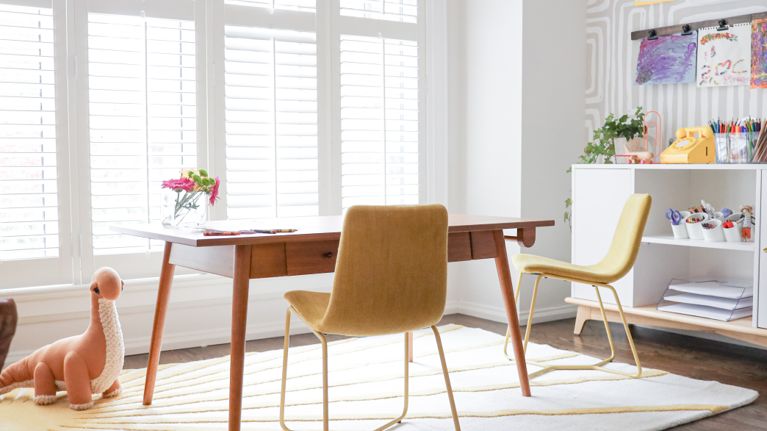 Two chairs sit by a wooden desk. In the background, a shelf full of art supplies.