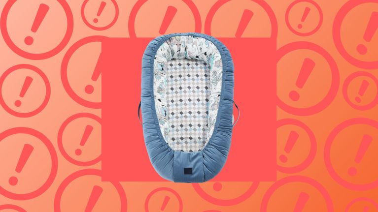 A soft, blue infant lounger with patterned interior padding is displayed against a bright red and orange background filled with exclamation mark icons, symbolizing caution. The product shown is the La Millou Baby Best Nest, which has been recalled in Canada due to potential risks of falls, entrapment, and strangulation.