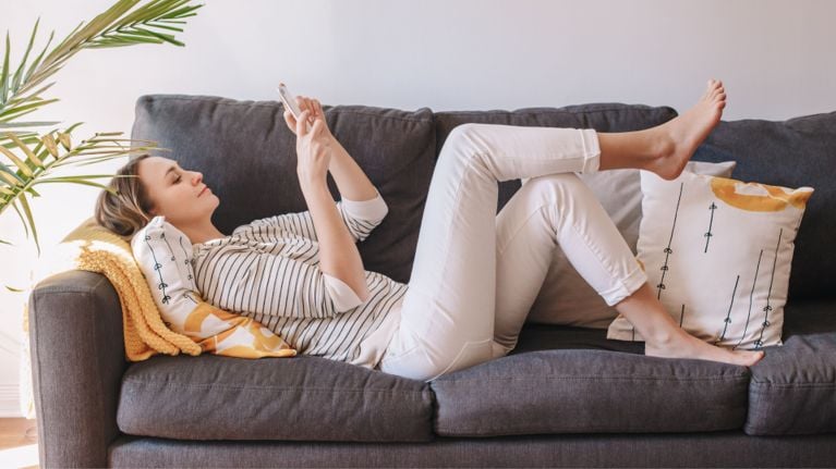 A relaxed woman lying on a dark grey couch, smiling while scrolling her phone. She’s barefoot, dressed in a striped shirt and white pants, surrounded by bright pillows and a yellow knit blanket, enjoying a quiet moment alone at home.