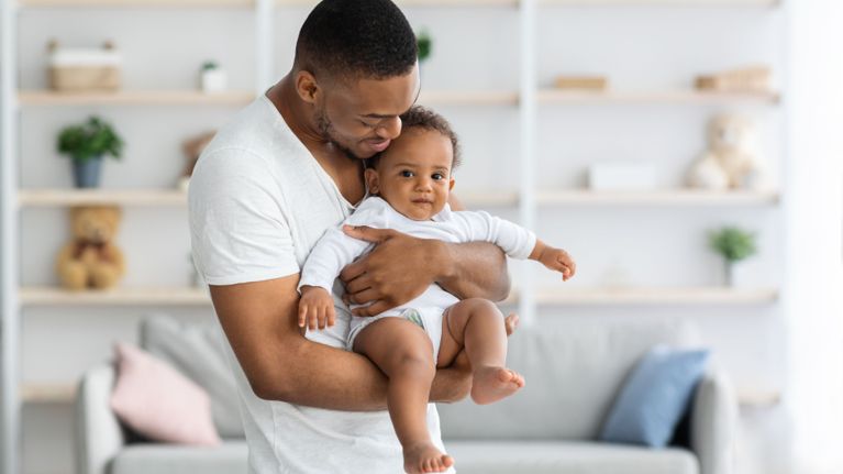 A man wearing a white tshirt holds a baby in a white, long-sleeved onesie. He smiles down at the baby.