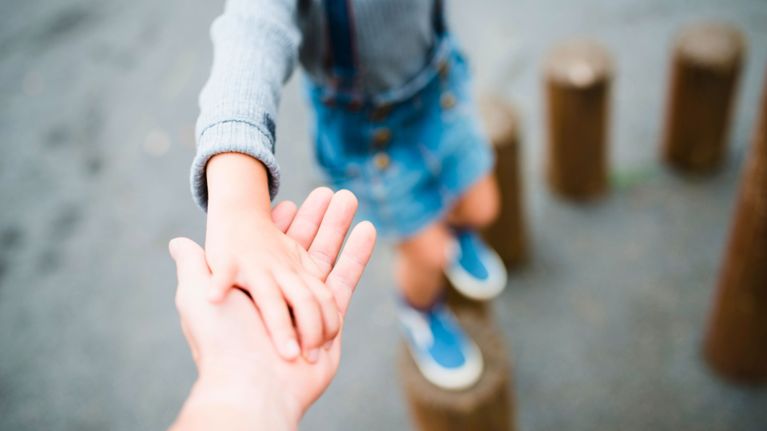 Close-up of an adult hand holding a child’s hand for support, as the child balances on wooden stumps in a playground. The image captures a tender moment of guidance and connection.