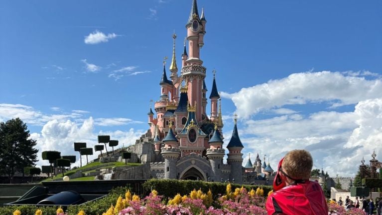 A young child dressed in a red superhero costume admires the iconic pink and blue Sleeping Beauty Castle at Disneyland Paris, surrounded by blooming flowers and topiary trees under a clear blue sky.
