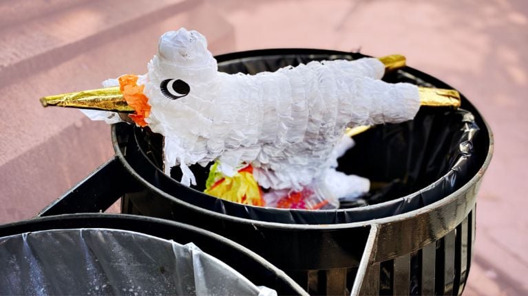 A torn white piñata with gold accents lying discarded in a black trash can, symbolizing waste and the fleeting nature of birthday party excess.
