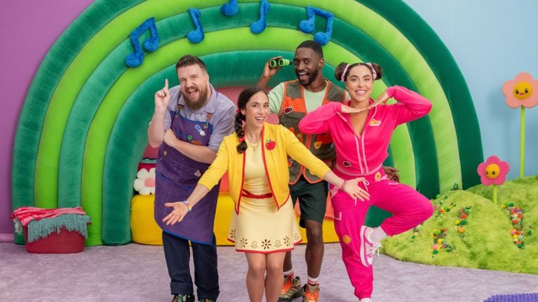 Ms. Appleberry, played by Allie Rivera Quiñonez, smiles brightly while holding a large plush character shaped like a watermelon with a pink face, blue antennae, and a ladybug, in front of a colorful rainbow arch with music notes in The Melon Patch set.