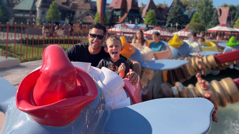 A smiling father and excited son enjoy a ride on Dumbo the Flying Elephant at Disneyland Paris, with other families riding in the background on a sunny day.