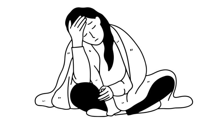 A woman sits on the floor wrapped in a blanket, holding her forehead with one hand and her knee with the other, appearing fatigued and unwell.