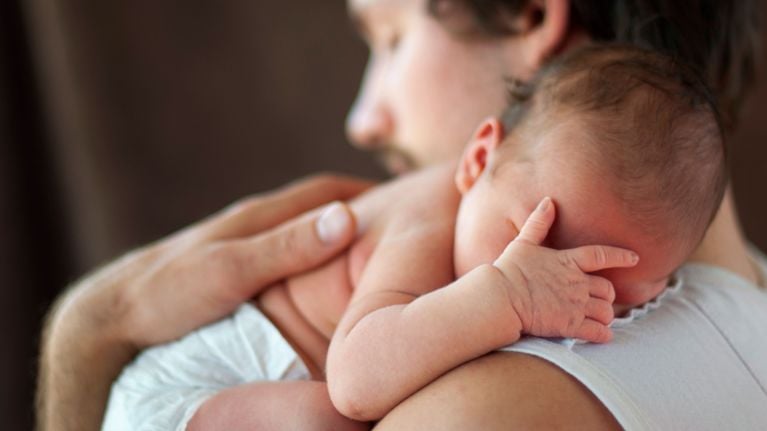 Newborn baby resting on a parent’s shoulder, covering part of their face with their tiny hand.