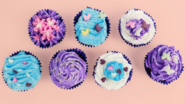 A row of brightly decorated cupcakes in purple, blue, and white icing with heart and butterfly sprinkles, set against a pastel pink background.