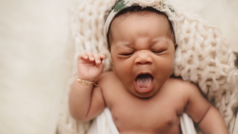 Baby wearing a headband and bracelet yawning while wrapped in a soft blanket.