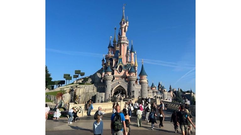 Crowds of visitors gather in front of the Sleeping Beauty Castle at Disneyland Paris, with clear blue skies and manicured topiary trees creating a picture-perfect scene.