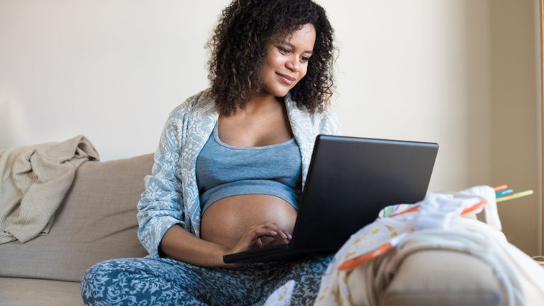 Pregnant person sitting comfortably on a couch in loungewear, using a laptop with baby clothes and essentials nearby.