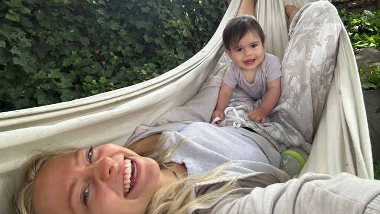 A woman smiles at the camera while lying in a hammock, with a happy baby sitting on her stomach. The background is lush with green ivy leaves.