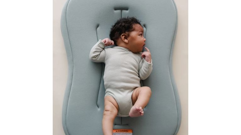 A baby in a sage green onesie lies on a blue-grey contoured baby pad, looking off to the side with one leg extended.