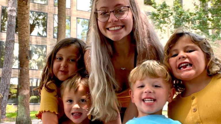 A smiling woman with long, light hair and glasses poses closely with four young children, all smiling at the camera, with palm trees and buildings behind them.