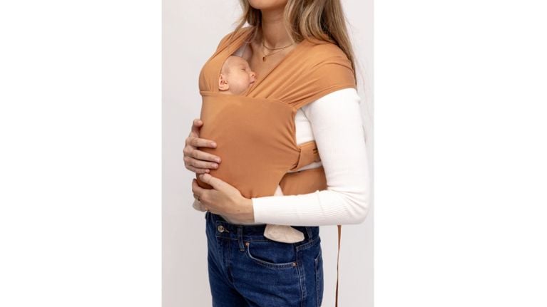 A caramel-colored wrap-style baby carrier worn by a woman holding a sleeping newborn against her chest.