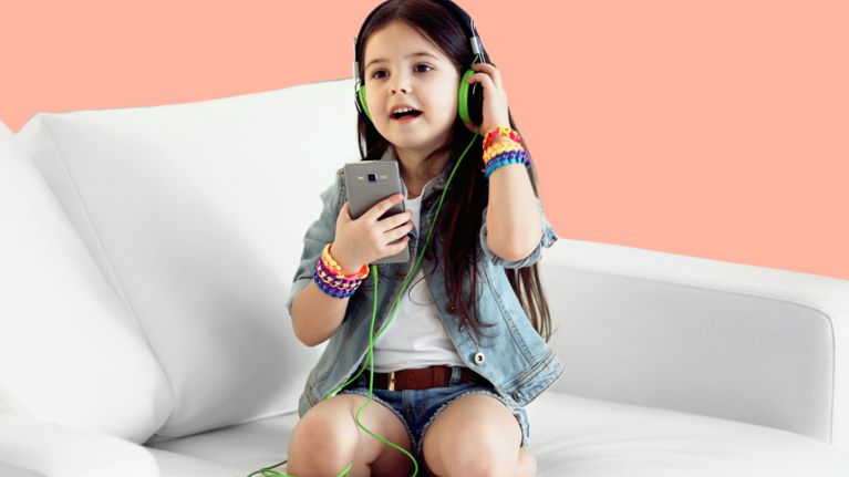 Young girl sitting on a white couch, wearing green headphones and colorful bracelets, holding a smartphone while singing along to music.