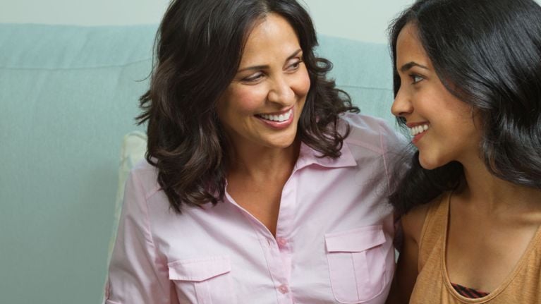 Mother and adult daughter sitting on a couch, smiling warmly at each other and sharing a close, relaxed moment, symbolizing a supportive and evolving parent–child relationship.