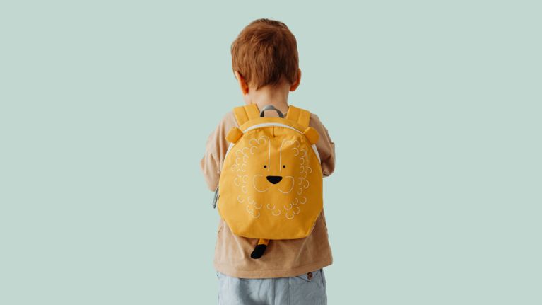 A young child with short light-brown hair, seen from behind, wearing a tan shirt and a bright yellow backpack with a cute lion face design, standing against a pale green background.