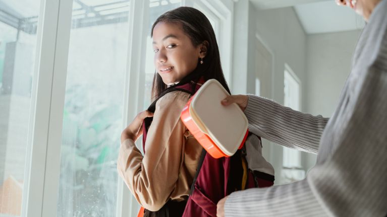 A smiling school-aged girl wearing a backpack looks over her shoulder as an adult places a lunchbox into her bag, standing by a bright window.