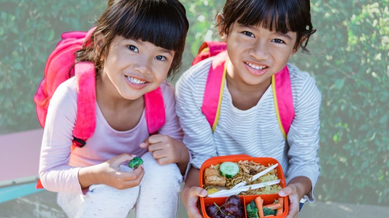 Two smiling young girls with pink backpacks sit outdoors, one holding a bright orange lunch container filled with noodles, rice, vegetables, and grapes, while the other holds a piece of broccoli.