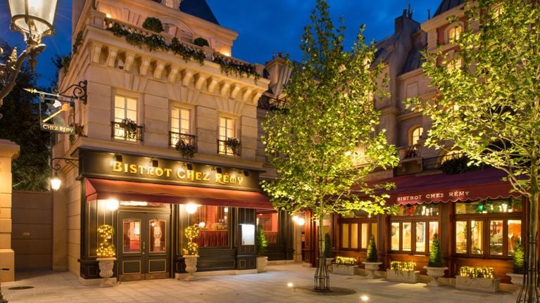 Exterior view of Bistrot Chez Rémy at Disneyland Paris, styled like a charming Parisian street café with warm lights, flower boxes, red awnings, and glowing trees at night.