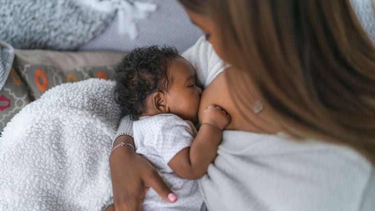 A mother breastfeeds her baby on a cozy bed. The baby is wearing a white polka dot onesie and is cradled in the mother’s arms, nursing peacefully against her chest.