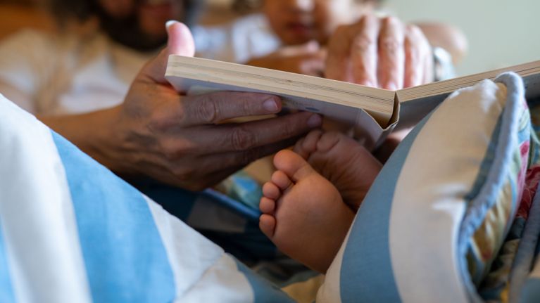 A close-up of an adult reading a book to a young child, with the child’s small bare feet resting on the adult’s lap under a blanket.