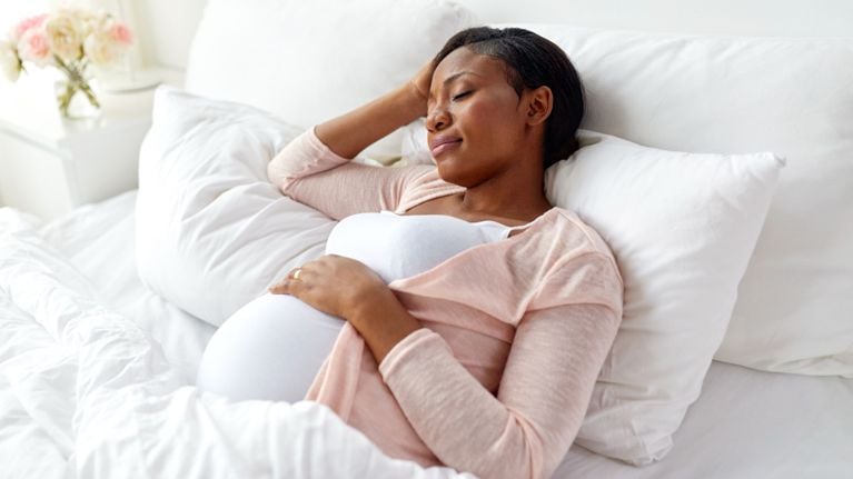 Pregnant woman resting peacefully on a bed with white linens, wearing a soft pink cardigan and cradling her belly.