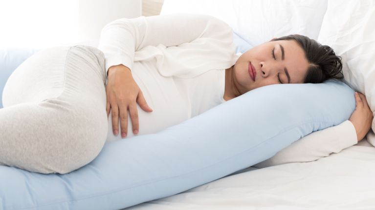 A pregnant woman lies on her side in bed, resting with her hand on her belly and supported by a large body pillow for comfort.