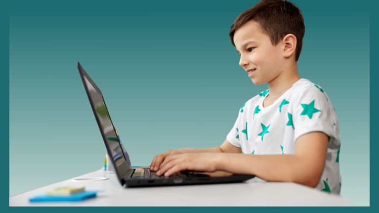 A boy in a white T-shirt with green stars types on a laptop at a desk, smiling as he looks at the screen.
